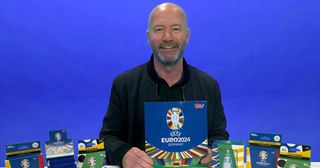 Alan Shearer in front of a blue background holding a Topps sticker book