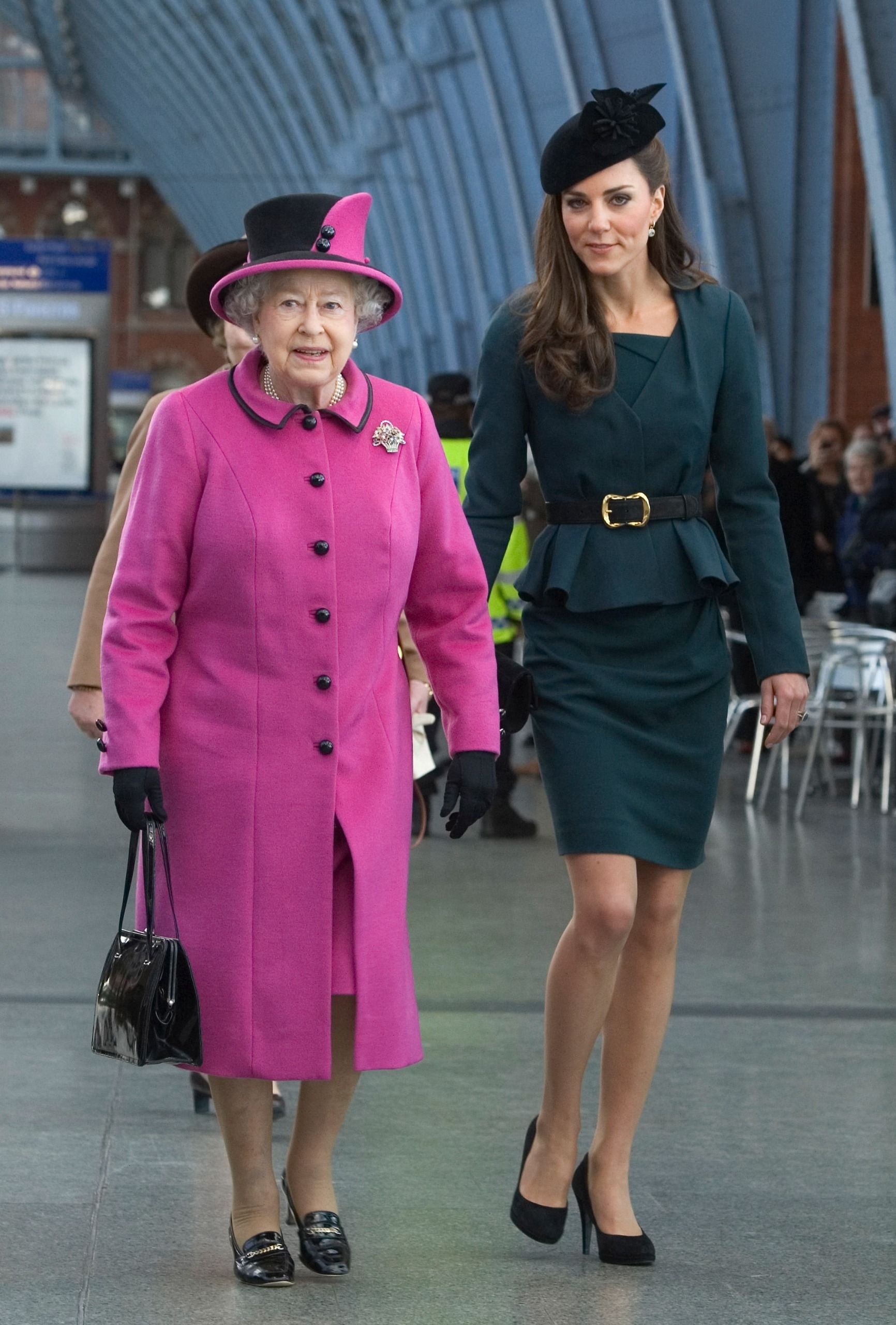 Queen Elizabeth in a pink coat walking next to Princess Kate, wearing a belted dress and black hat