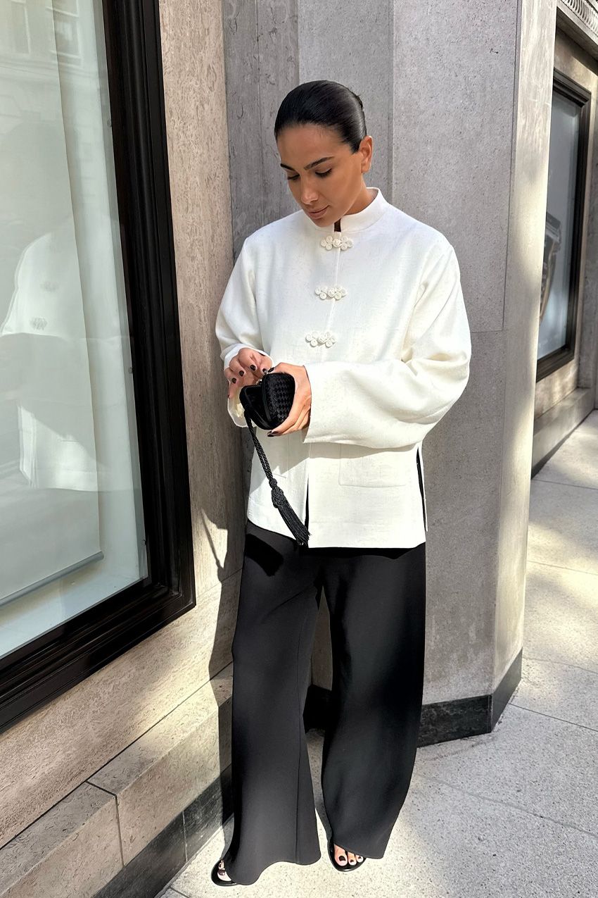 A woman standing outside wearing a white tang jacket styled with black wide leg trousers, black sandals, and a black clutch bag.