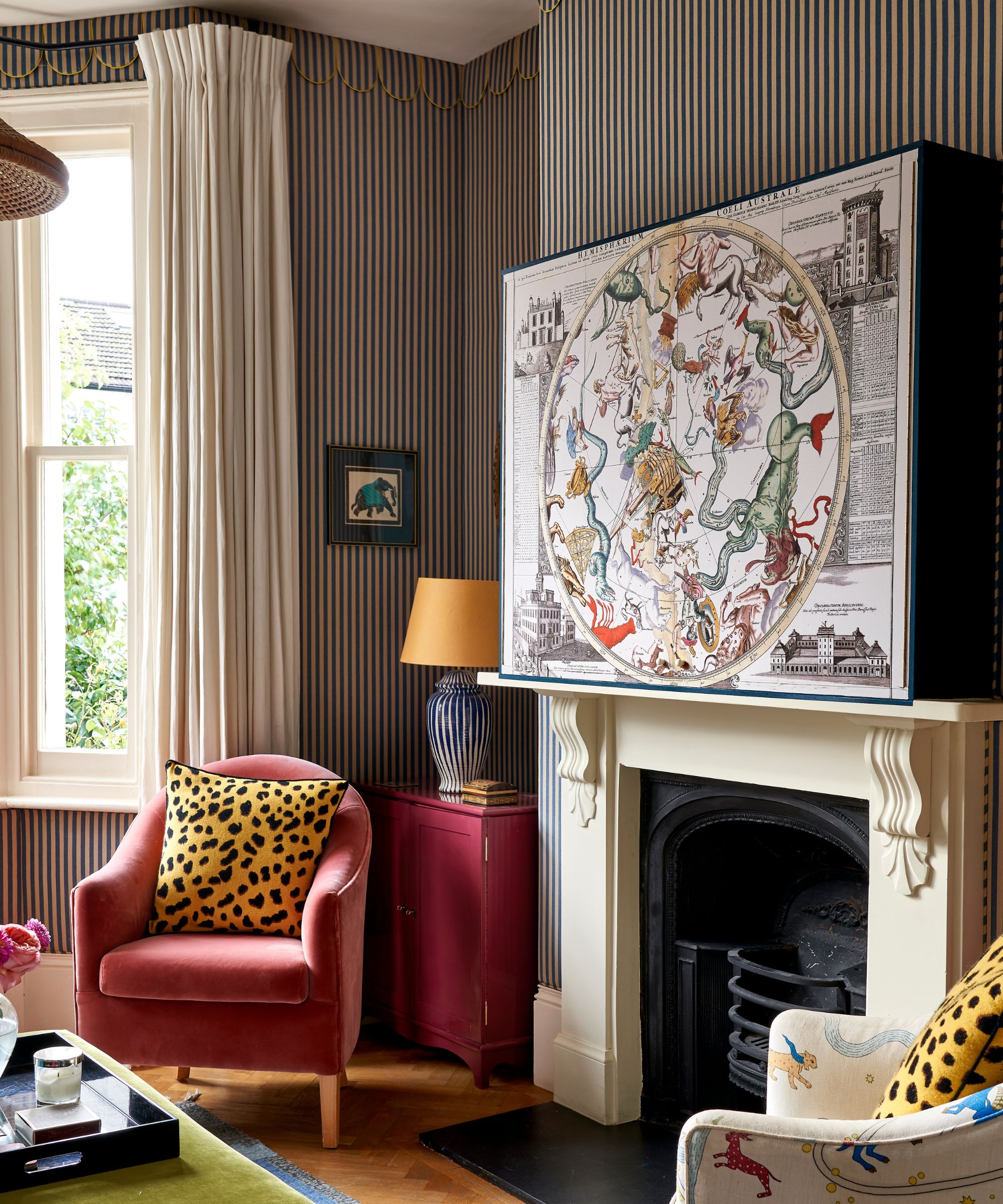 a striped living room with traditional fireplace, large artwork box hiding a TV, a small pink armchair with a leopard print pillow and a series of eclectic side tables in the bay window and alcove