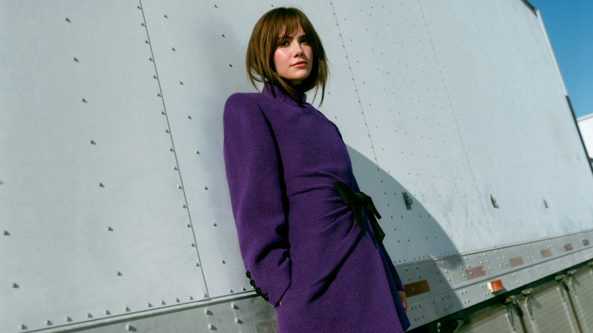 Emilia Jones for WWW's Ones to Watch story. The actress is leaning against a white semi truck wearing a purple coat with her hair tucked into the coat.