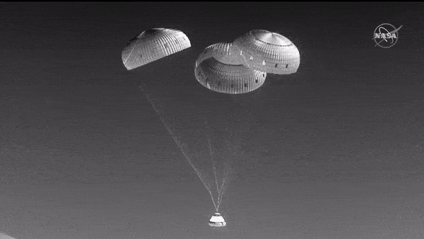 black and white video of a cone-shaped spacecraft touching down in the desert under 3 parachutes