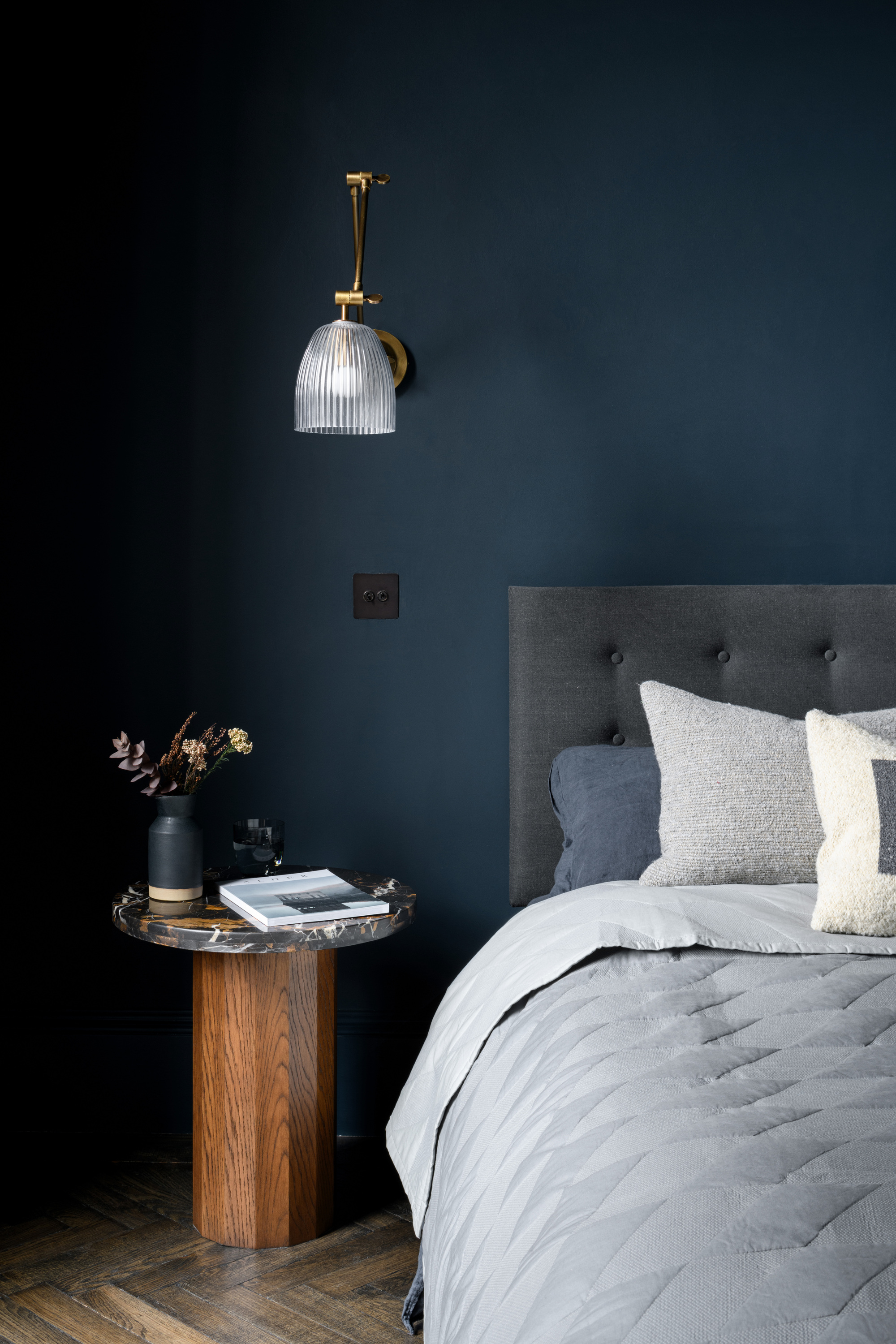 blue bedroom with gray headboard and bedding, timber and marble round side table on parquetry floor, and a wall sconce hanging overhead