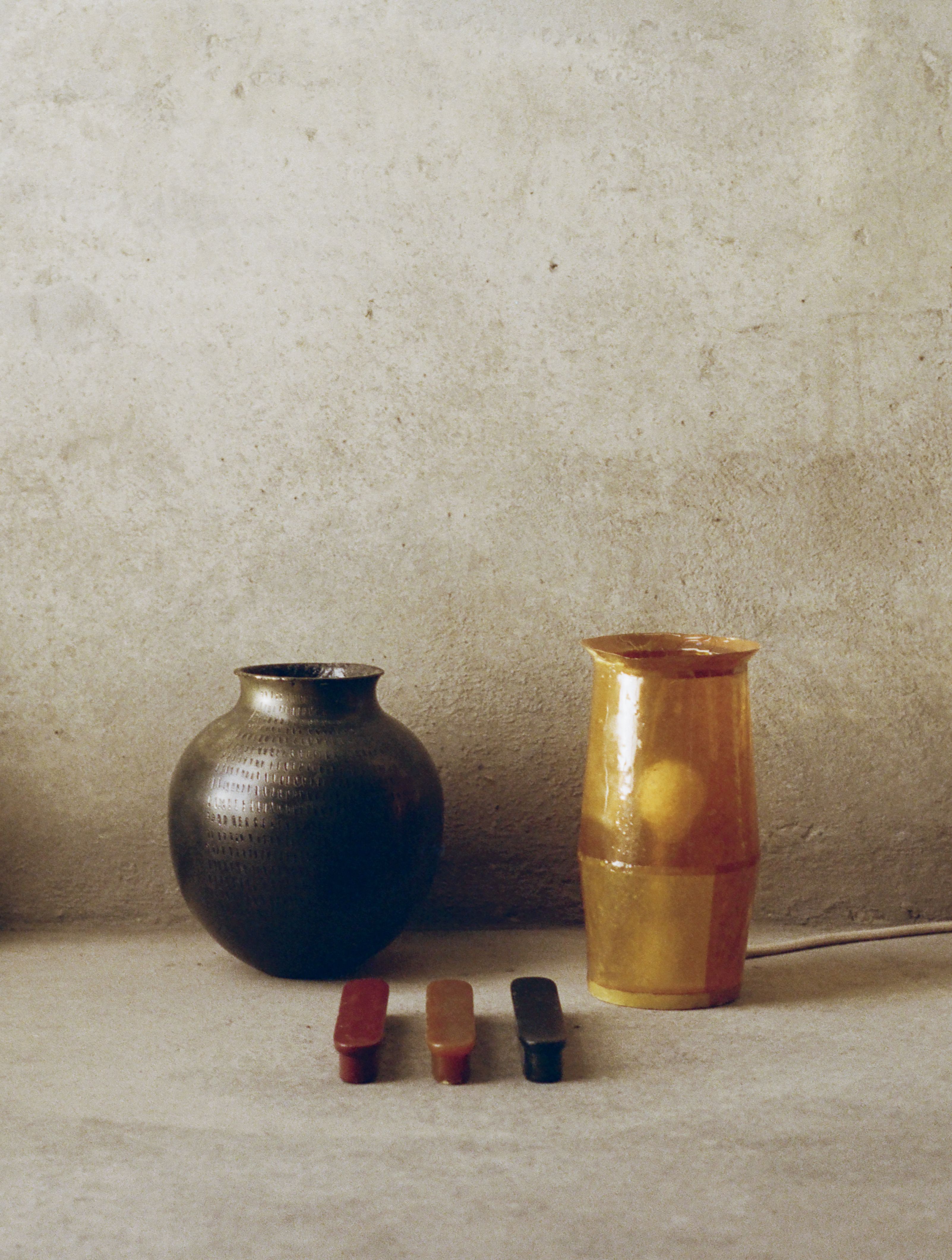 A woman throwing ceramic at a wheel and two vases, one in black earthware, one in golden resin, sitting next to three home accessories in red, gold, and black, against a beige background.