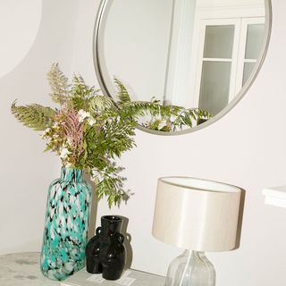 A vase containing leafy green stems sits on a side table with a lamp under a round mirror