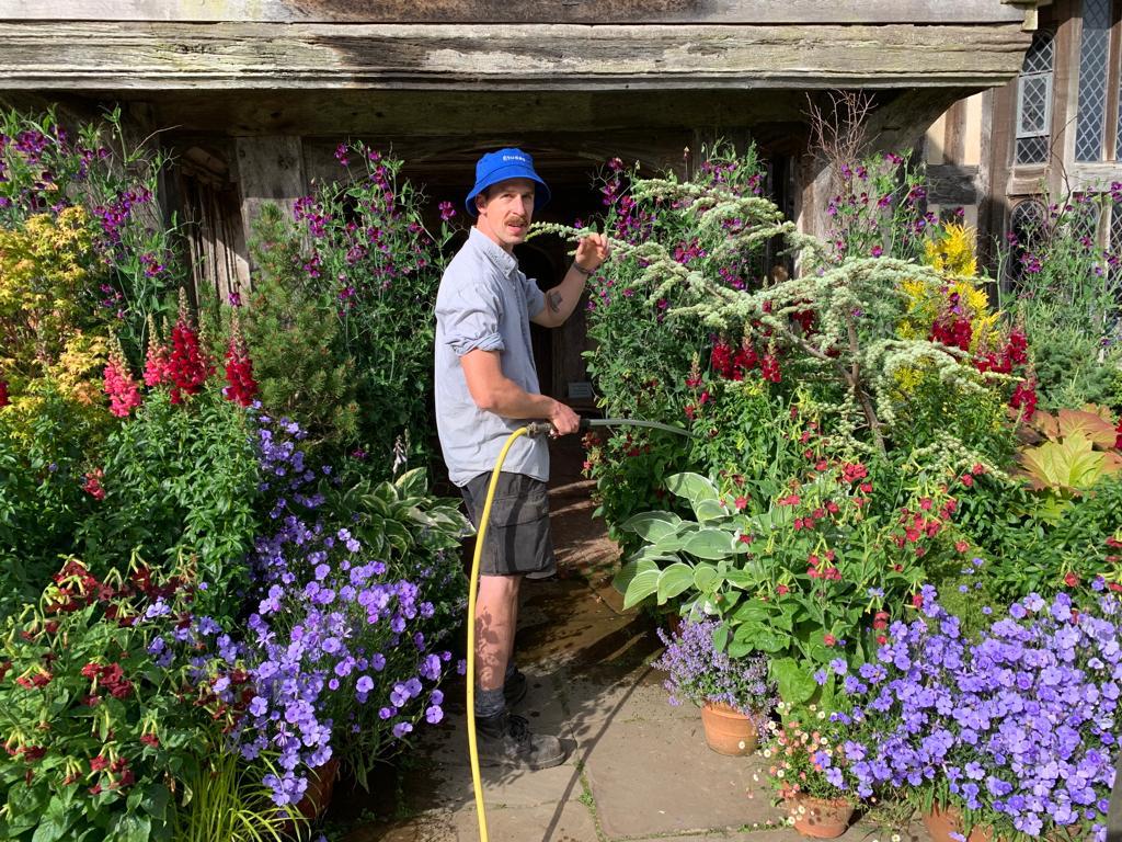 A young gardener waters plants in a blue short sleeved shirt, bucket hat in blue, and black shorts.