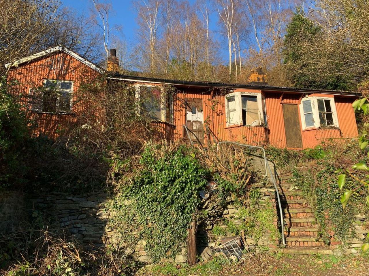 Steps leading up to a bungalow with smashed windows and overgrown garden