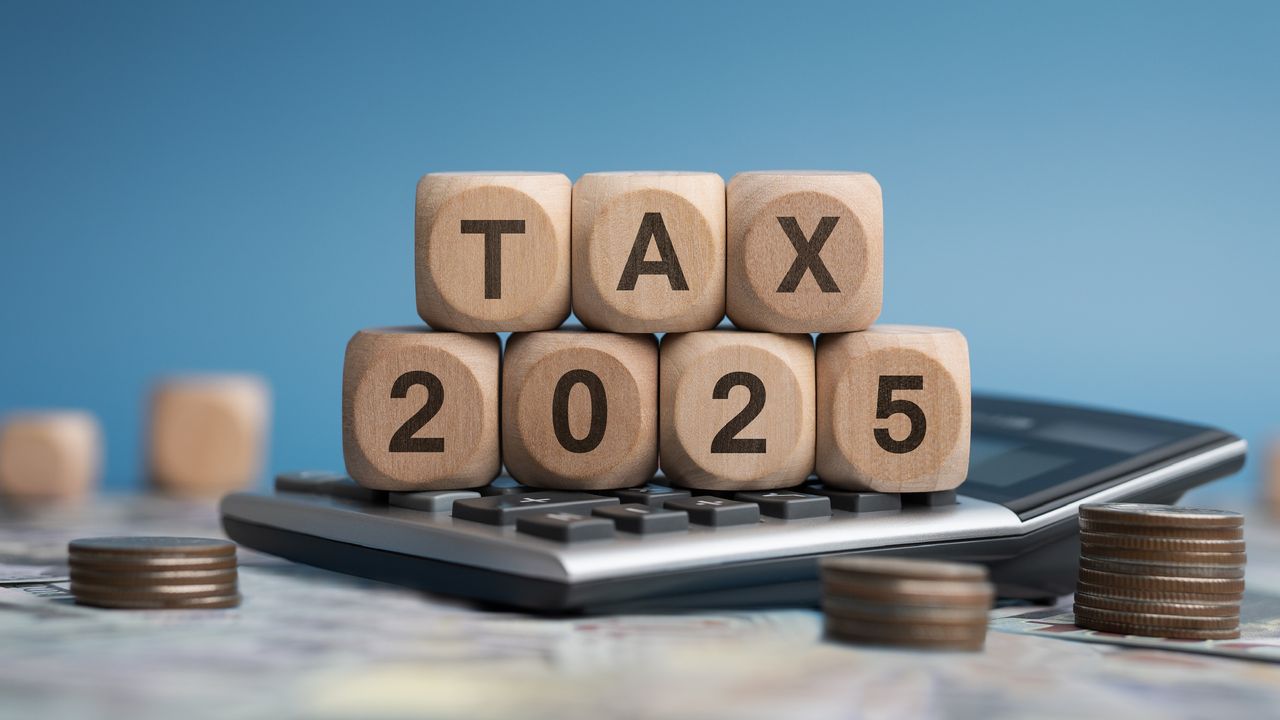 Wooden blocks spell out &quot;tax 2025&quot; on top of a calculator, surrounded by small stacks of coins.