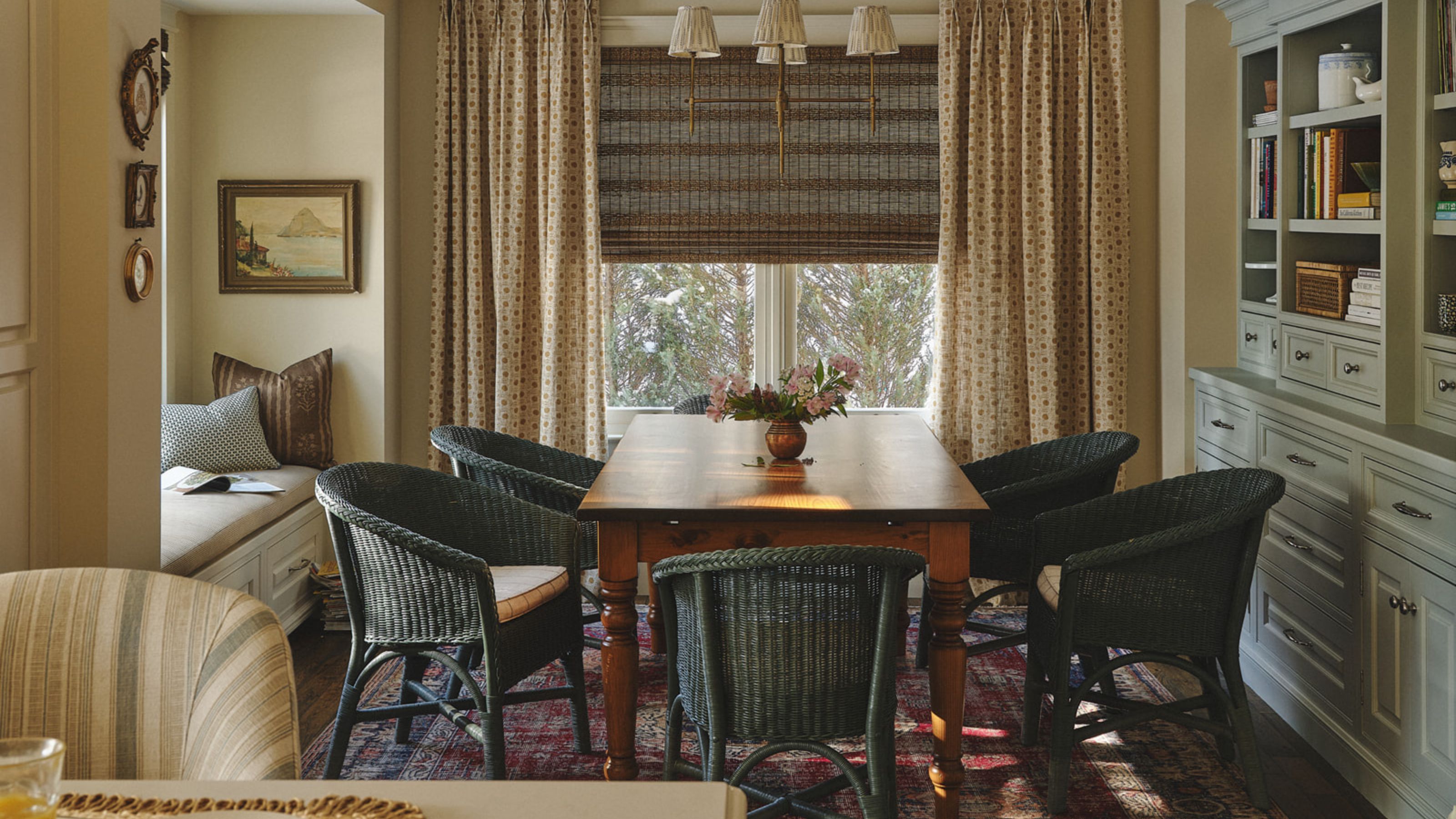 a warm soft yellow kitchen dining area with a window seat, antique farmhouse table and wicker chairs, vintage rug, bamboo blinds and a painted hutch