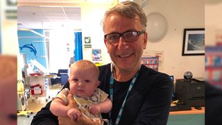 photo of an older white man with glasses holding a blonde baby wearing a onesie in the lobby of a hospital