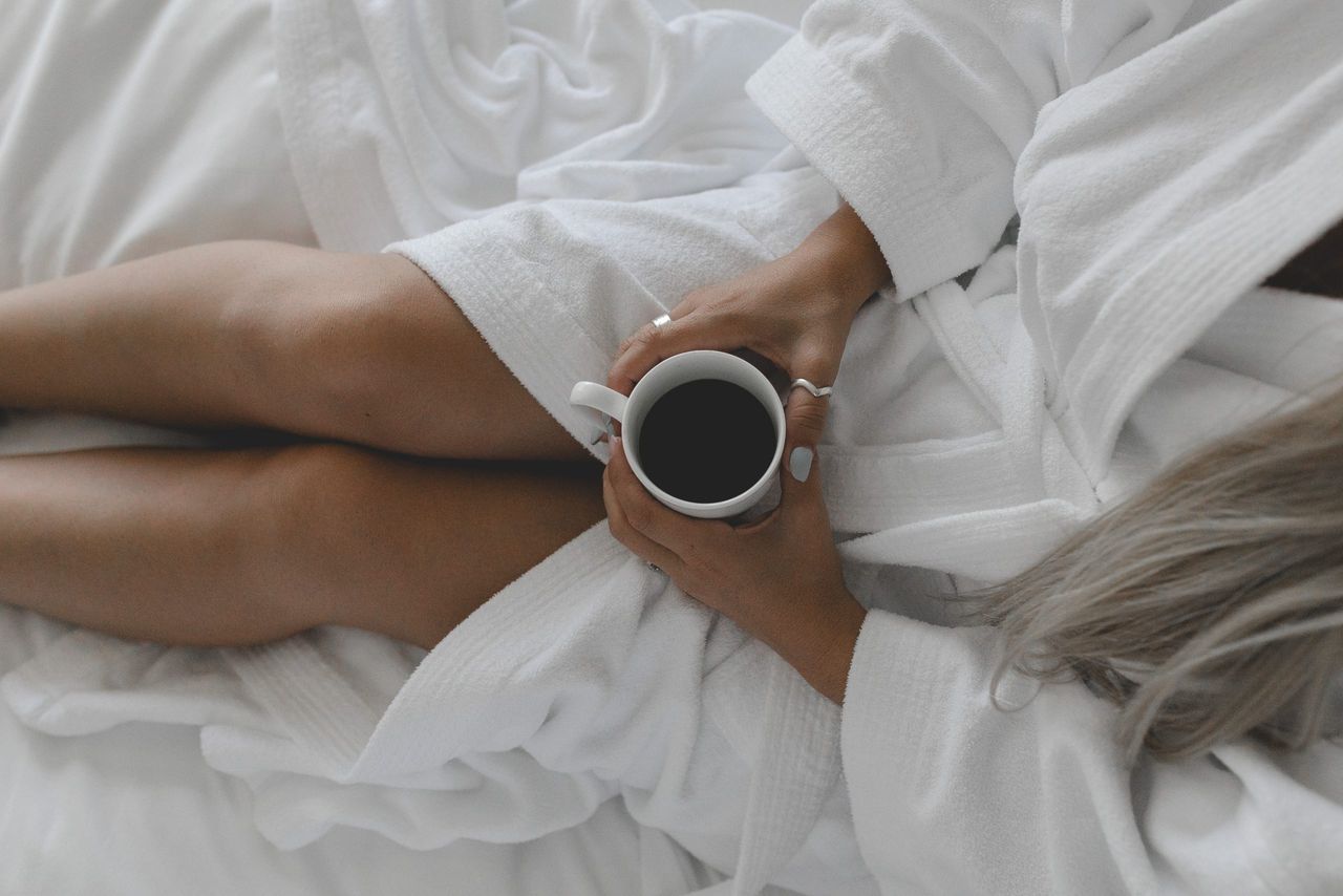 bedroom decor ideas: A young tanned woman sitting on a white bed in a white robe holding a coffee cup shot from above.