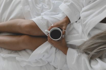 bedroom decor ideas: A young tanned woman sitting on a white bed in a white robe holding a coffee cup shot from above.