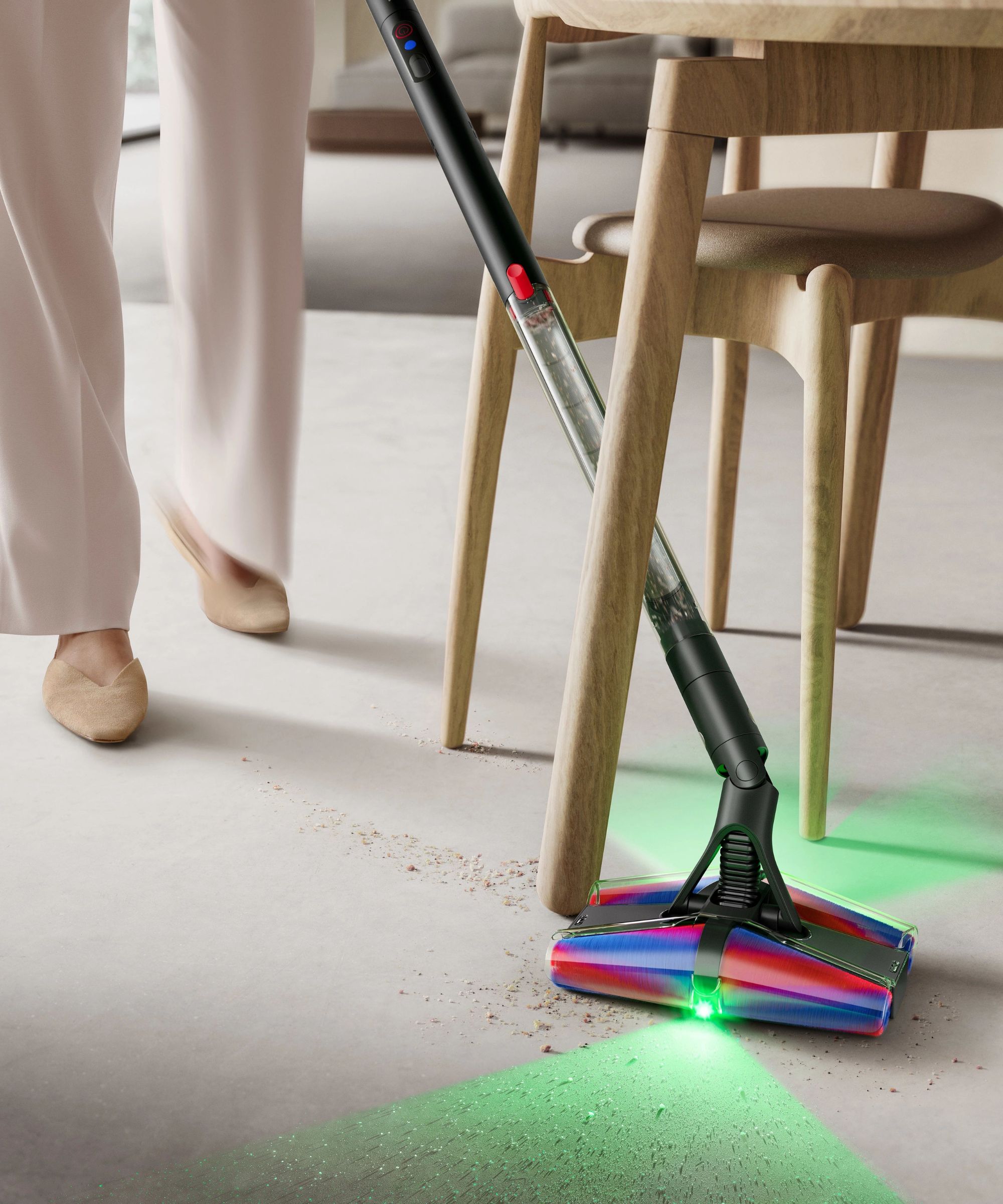 Dyson PencilVac cleaning small debris between wooden table and chair legs, using green light to illuminate dirt on floor. Pictured behind are person's legs and a cream living room and home.