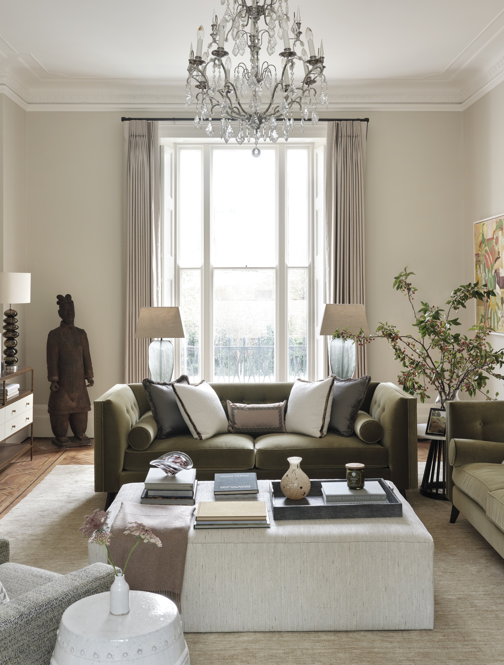 traditional green and gray living room with chandelier, fabric ottoman styled with tray, books, and small vases, a green sofa with lots of throw pillows, two lamps on a console table behind the sofa, a statue in the corner, a side table with a vase with large branches, and a window with beige curtains