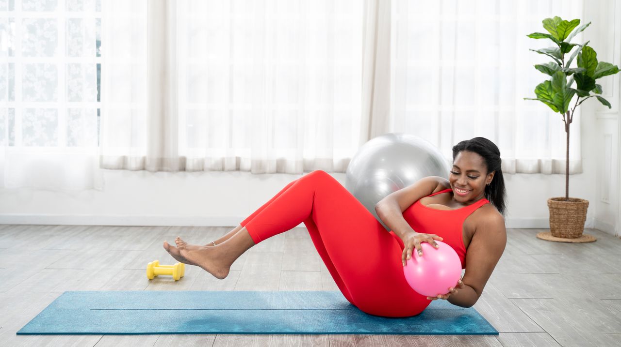 A woman in leggings and a sports top performs a crunch with a Pilates ball on an exercise mat. Her buttocks are on the floor, knees bent, feet elevated, and she holds the ball in her hands as she twists to the side. Behind her we see a large window and leafy platnt.