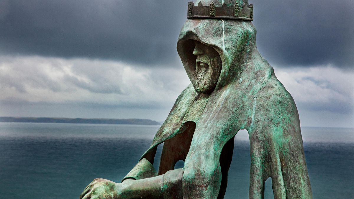 A turquoise rusted metal statue shows a man wearing a bronze crown over a hooded cloak with his hands in front of him and a sea shore behind him.