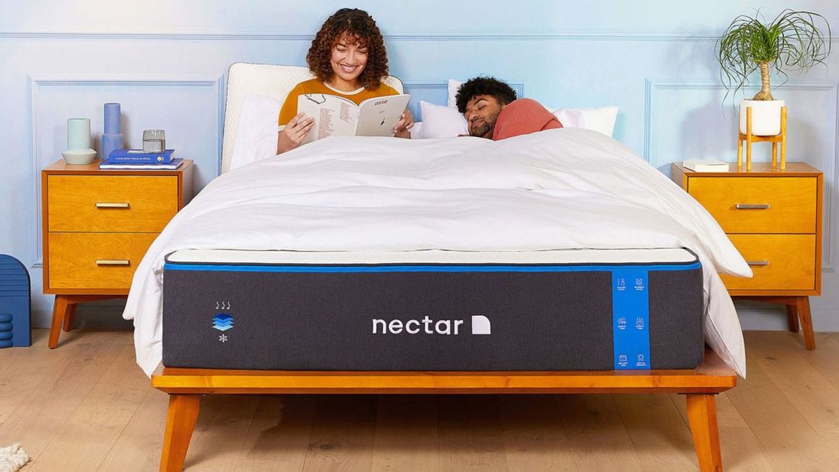 A woman smiles while reading next to her husband who is asleep on his side on the Nectar Memory Foam Mattress