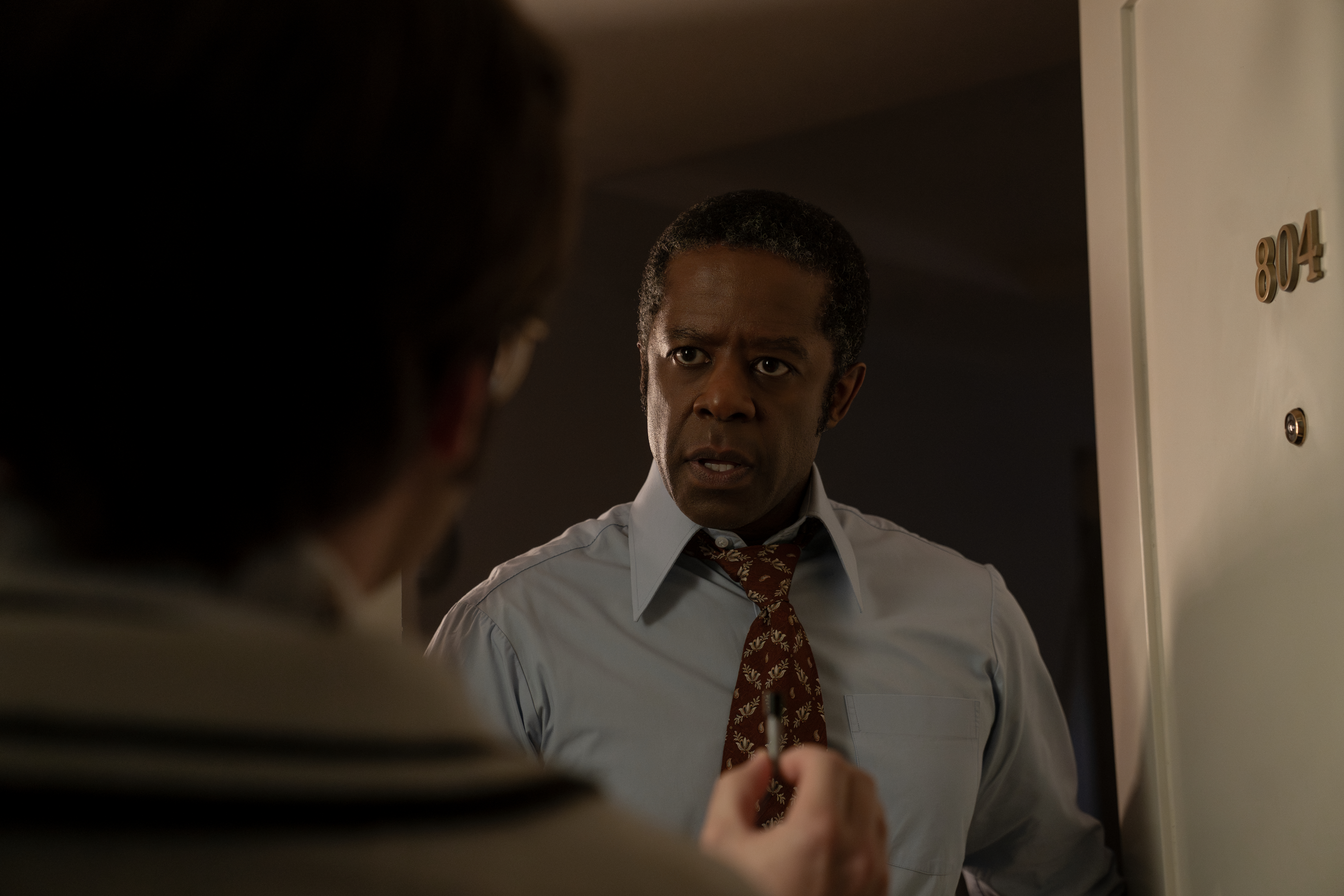 A series of film stills and photographs from a 1970s-inspired set featuring vibrantly decorated interiors with layered furniture and warm lighting, along with characters dressed in vintage clothing whether indoors or at parks, in cards, or at markets outdoors.