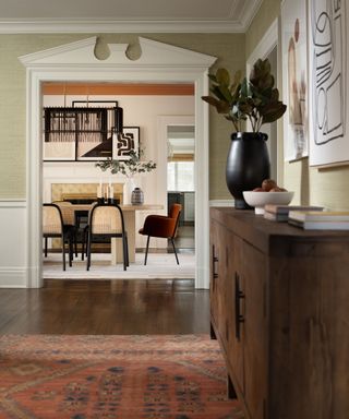 wallpapered entryway hallway with a terracotta vintage rug with wooden credenza and a view into the dining room with a doorway with decorative molding