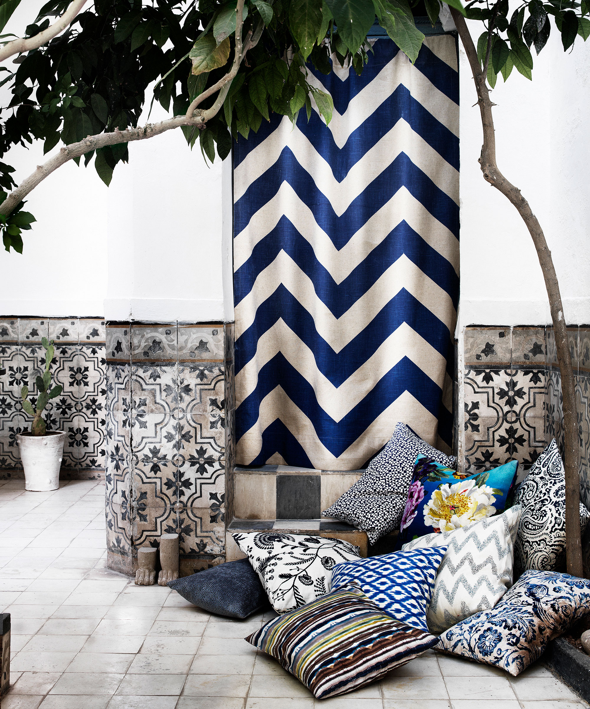 Colorful fabrics and cushions clustered in the corner of a Morrocan inspired courtyard garden idea with faded white and patterned tiles.
