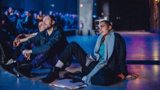 Three stylish men sat on the floor bathed in purple and blue light interestedly observing and listening to a speaker (not shown)