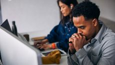 Visibly stressed male office worker looking at desktop computer screen with clenched fists placed under chin, while female colleague works on laptop in background.