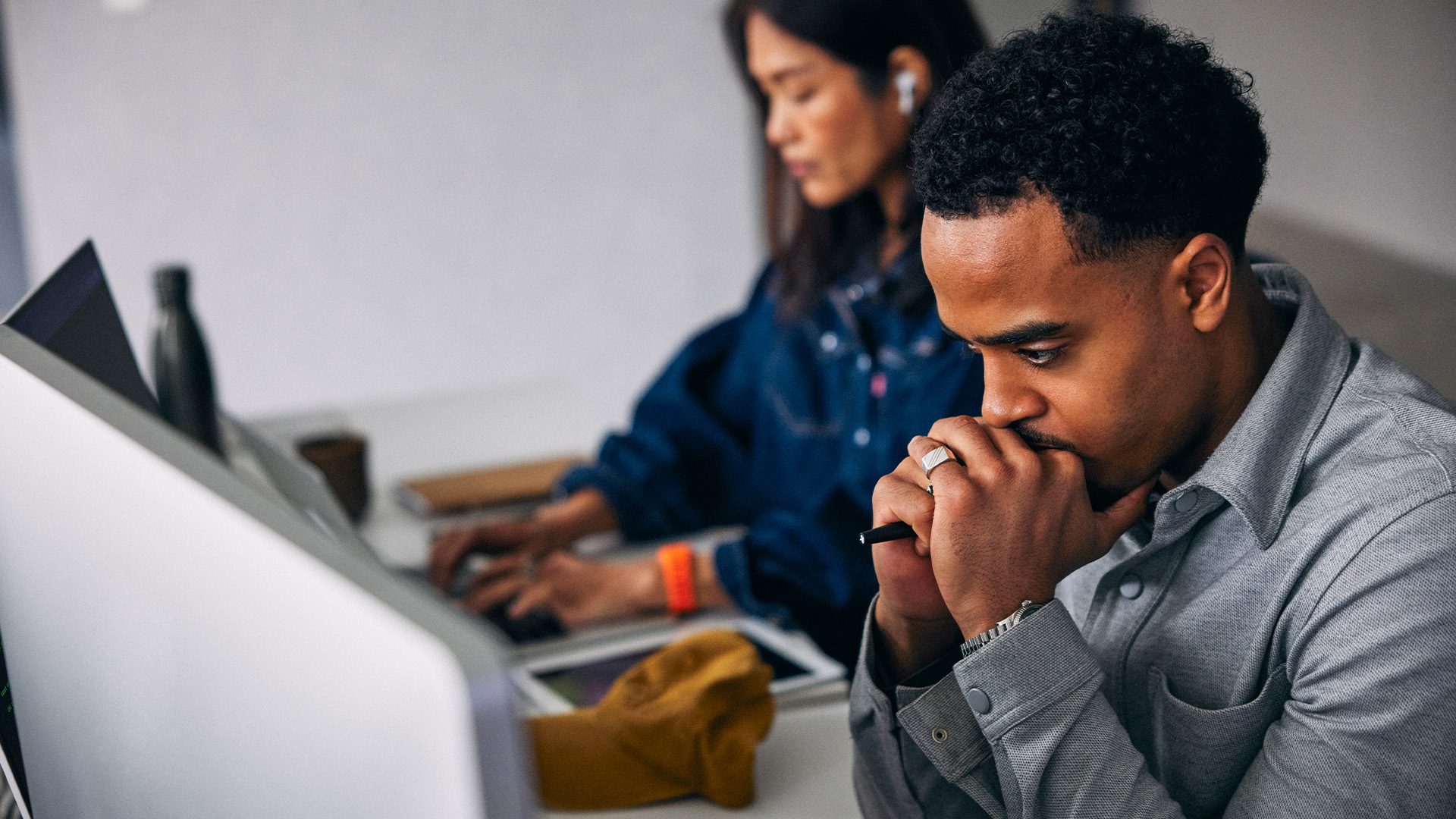 Visibly stressed male office worker looking at desktop computer screen with clenched fists placed under chin, while female colleague works on laptop in background.
