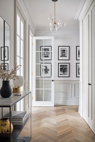 Hallway with wooden flooring, pale grey part panelled walls with framed black and white photographs, metal and marble topped console table and glass doors