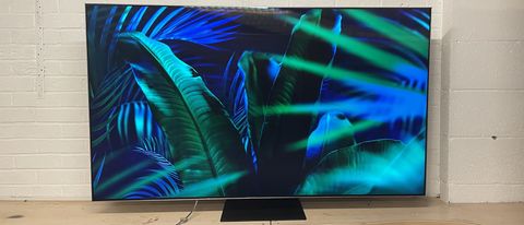 En Samsung QN95B QLED TV på en TV-bänk och visar en bakgrundsbild av tropiska växter som skiftar i mörkblått och mörkgrönt.
