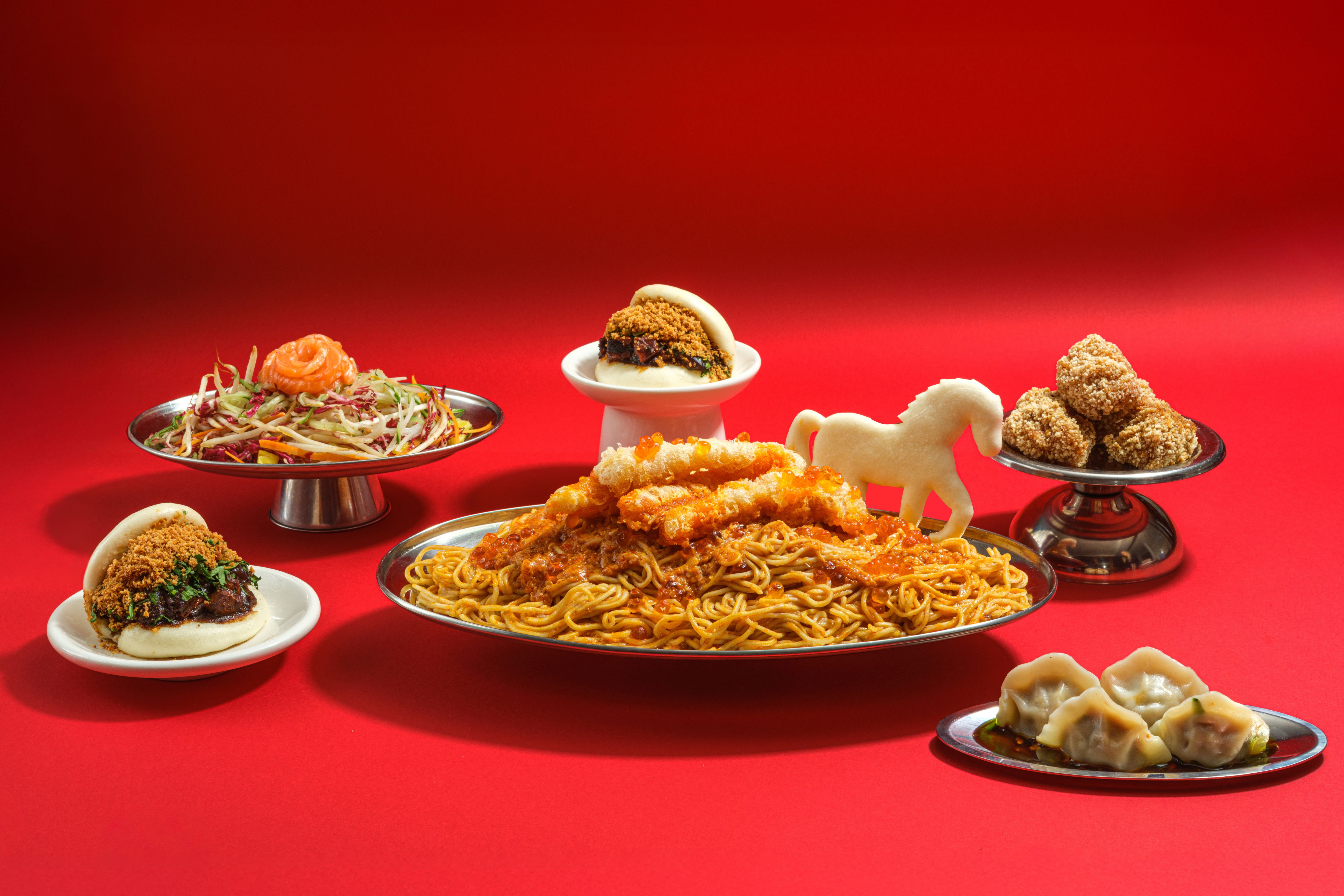 A styled shot of a series of Chinese food plates, placed inside silver as well as white ceramic serveware and set against a bright red background, with the central dish boasting a horse-shaped piece of dough.