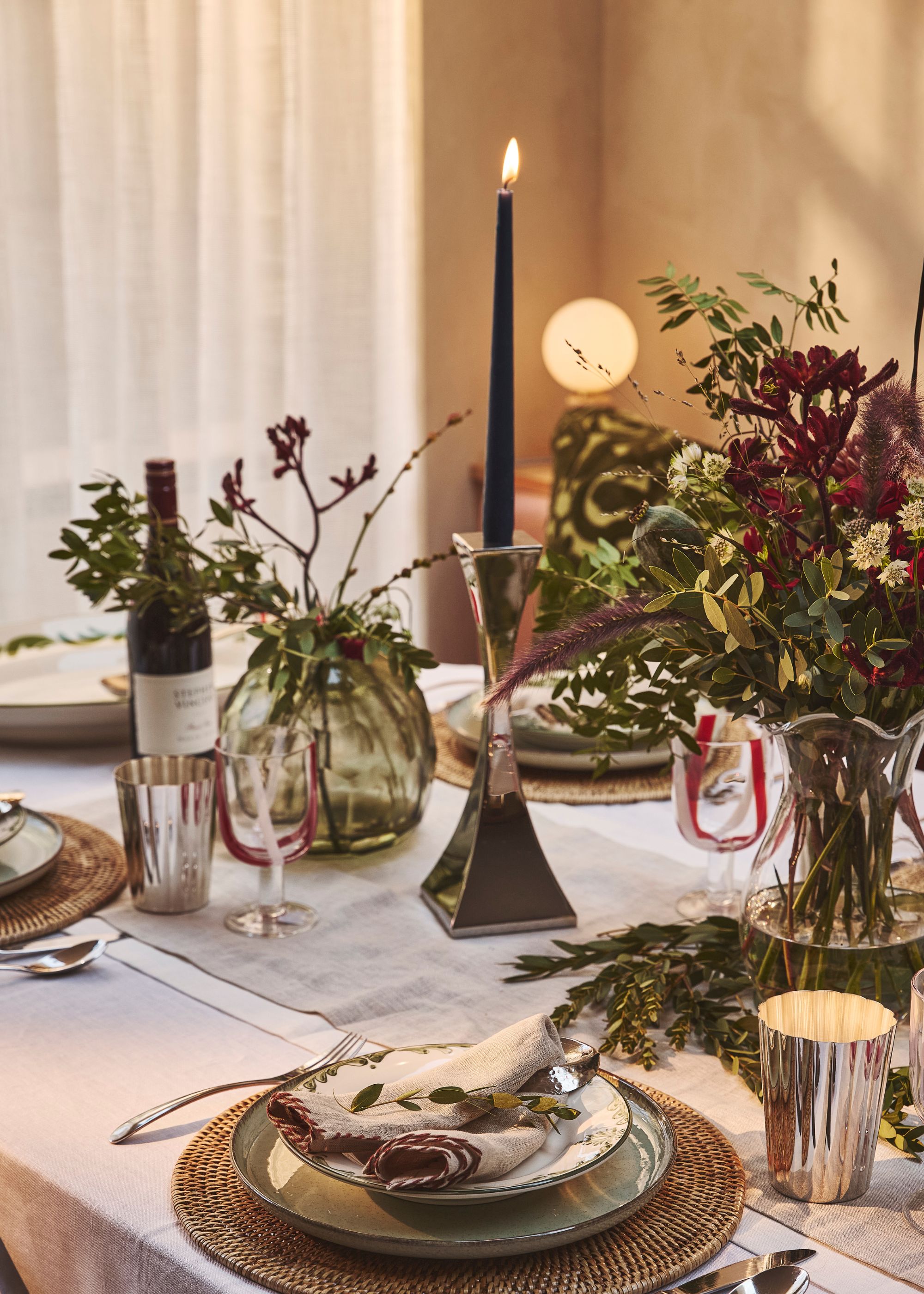A lit candle on a dining table next to bright foliage and flowers in a vase. On the table is also tableware such as a runner, plates, utensils, napkins and glassware