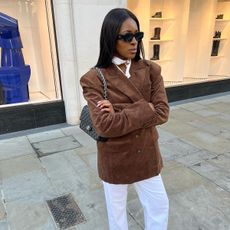 British influencer @georgia_3.0 wearing a brown suede blazer, white jeans, Chanel classic flap bag, and black sunglasses