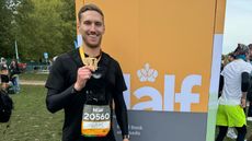 Man stands in front of a orange sign holding medal. He is wearing a black top with a race number pinned to it