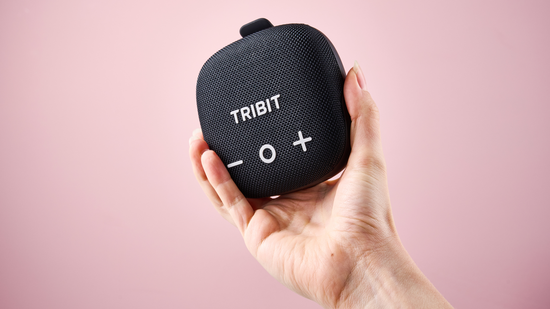 A woman's hand holding up the Tribit Stormbox Micro 3 in front of a pink background.