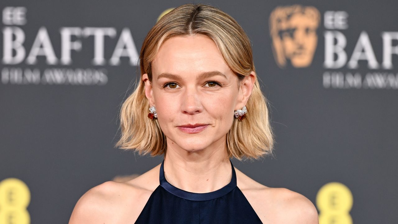 Carey Mulligan is pictured with a wavy bob, with the front strands tucked behind her ears, and wearing silver earrings and a navy Prada dress at the 2026 EE BAFTA Film Awards at The Royal Festival Hall on February 22, 2026 in London, England. 