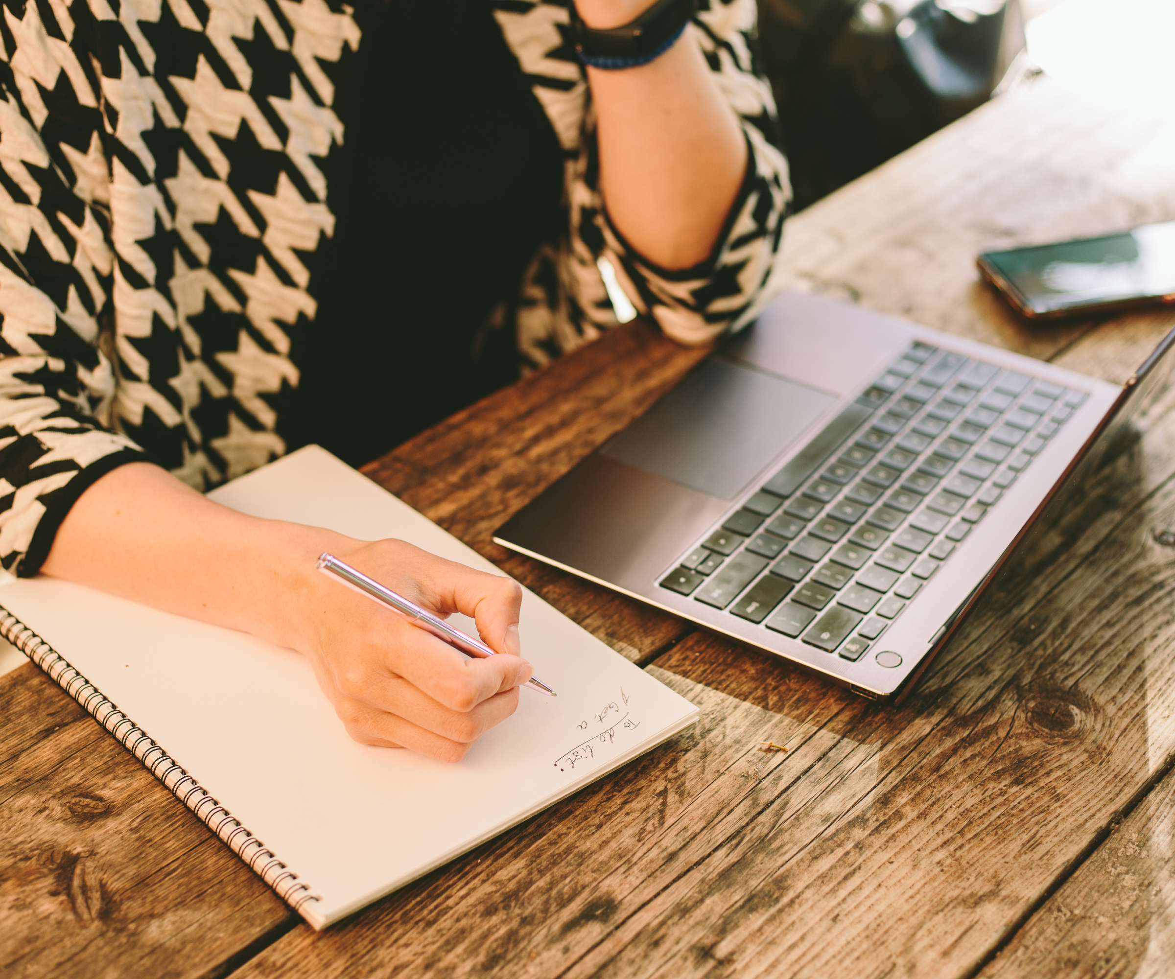 female sat at wooden table with notebook, pen and laptop making notes