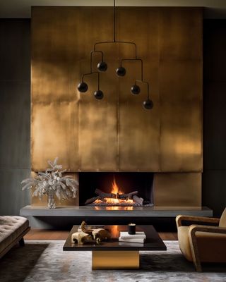 A dark living room with a grand gold metallic fireplace that stretches from floor to ceiling. There is a gold and black rectangular coffee table in front of the fireplace and an accent chair halfway in the frame