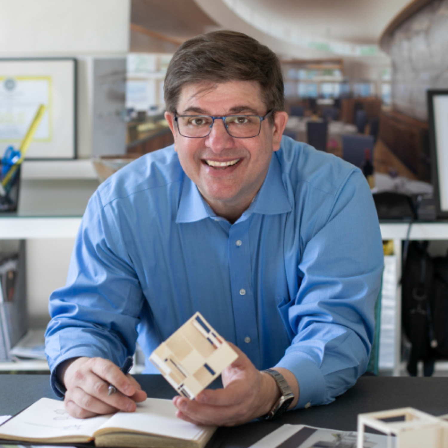 A smiling man in a blue shirt and glasses sitting by a desk