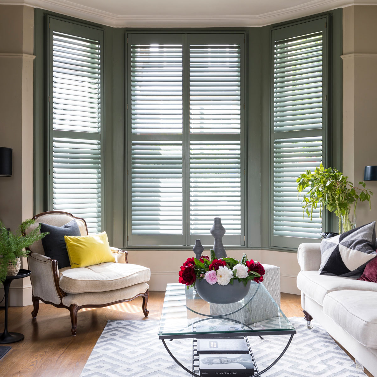 Bay windows with green full wooden shutters and cream fabric couch, armchair and area rug