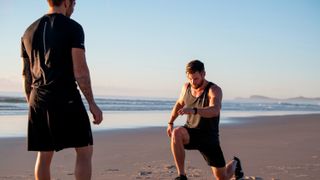 Chris Hemsworth performing lunge exercises on the beach, his trainer Luke Zocchi looks on