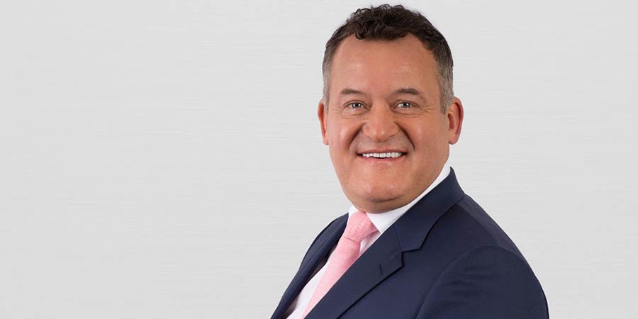 A headshot of Paul Burrell wearing a blue suit and pink tie