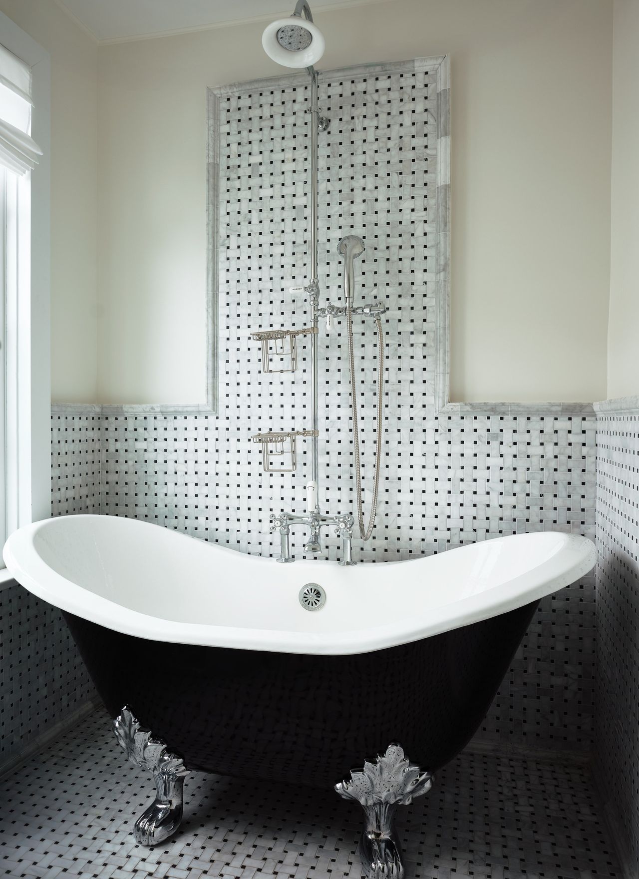 Image of a bathroom where the wall and flooring are covered in a gray basket weave tile. There is a stand alone tub that is black with silver feet.