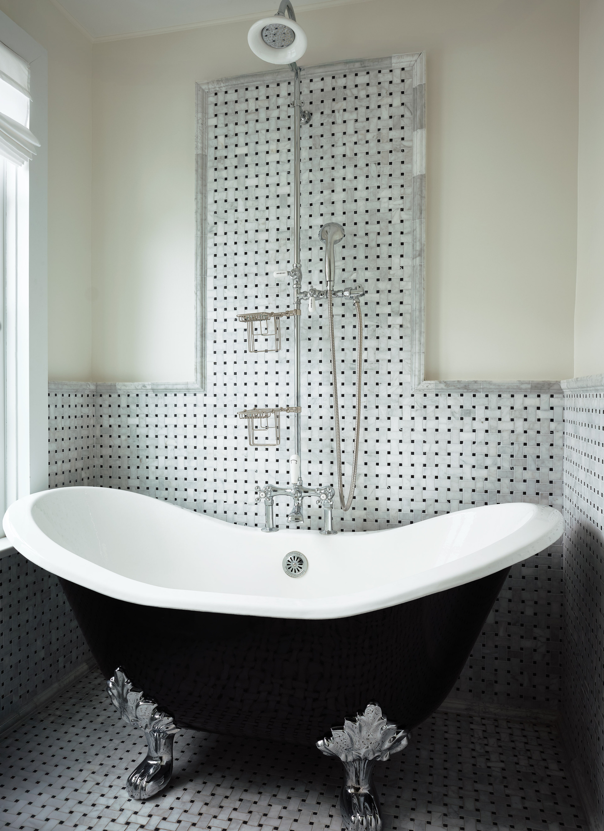 Image of a bathroom where the wall and flooring are covered in a gray basket weave tile. There is a stand alone tub that is black with silver feet.