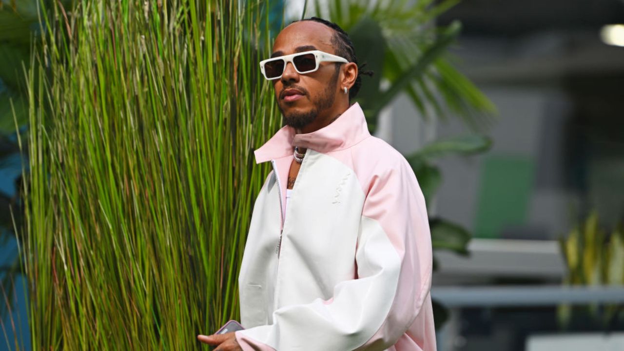 Lewis Hamilton&#039;s living room is so chic. Here is the Formula 1 star walking in fromt of a green grass hedge wearing white sunglasses and a pink and white jacket