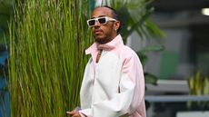 Lewis Hamilton's living room is so chic. Here is the Formula 1 star walking in fromt of a green grass hedge wearing white sunglasses and a pink and white jacket