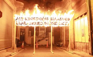 The words "ALL KINGDOMS SMASHED AND BURIED IN THE SKY" on fire