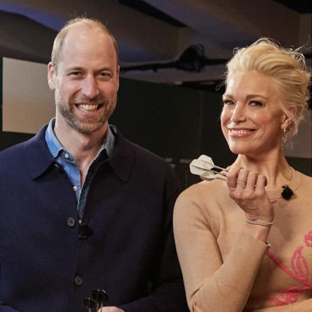 Prince William wears a navy sweater over a blue shirt as he poses Hannah Waddingham who has blonde hair, wears a taupe sweater, and is holding some darts