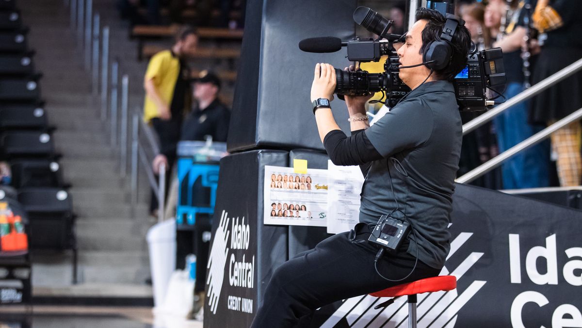 A cameraman sits courtside at a Big Sky basketball game recording with a JVC camera. 