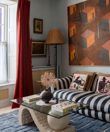 Living room with pale blue painted walls, large painting of houses in orange and red, a striped blue and white sofa, a glass and stone coffee table with decor on top and red curtains