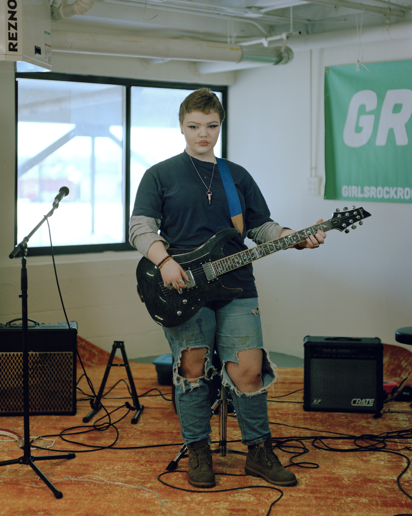 A girl with short hair and ripped jeans holds an electric guitar in a rehearsal room with amps, cables and a microphone stand